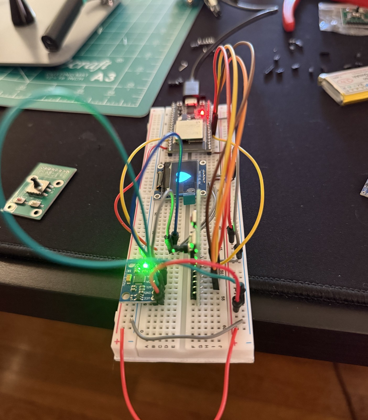 The badge on a breadboard, ESP32 and OLED wired up with jumpers, a cutting mat and a spare breakout board on the desk